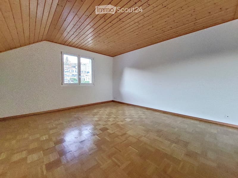 Empty room with wooden floor, white walls, and a slanted wooden ceiling. Two windows on one wall provide natural light.