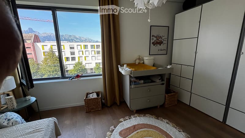 A well-lit bedroom with a bed, changing table, wardrobe, small table, and various toys. The room features large windows with a scenic mountain view and a cozy rug on the floor.