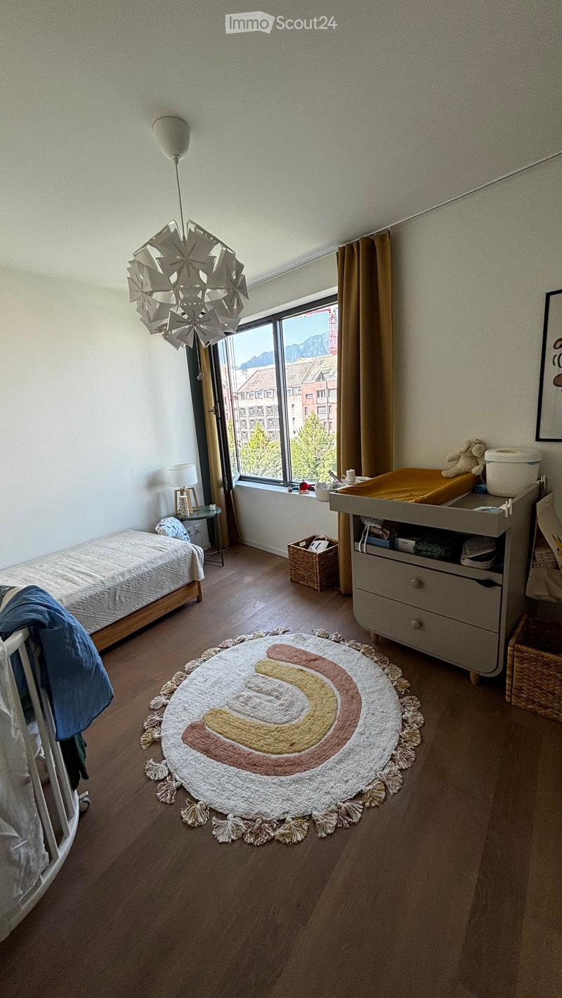 Brightly lit bedroom with wooden flooring, a single bed, a baby crib, a changing table, a decorative rug, and a large window offering a scenic view of mountains and buildings.