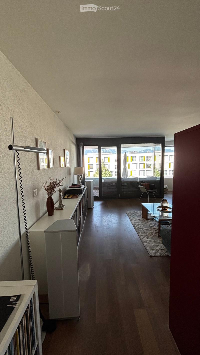 Modern living room with wooden flooring, large sliding glass doors leading to a balcony, a sofa, a coffee table, a table lamp, a vase with dried flowers, and a cabinet with picture frames.