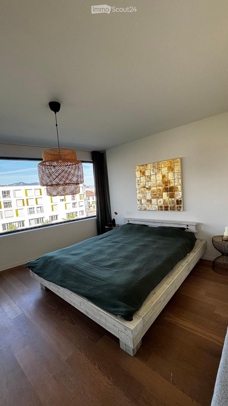 Modern bedroom with a wooden bed, dark green duvet, pendant light, nightstand, and wall art.