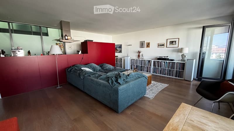 Modern living room with a grey sectional sofa, coffee table, floor lamp, and a sleek black chair. The room features wooden flooring, large windows, and a red accent wall with a bookshelf and decorative items.