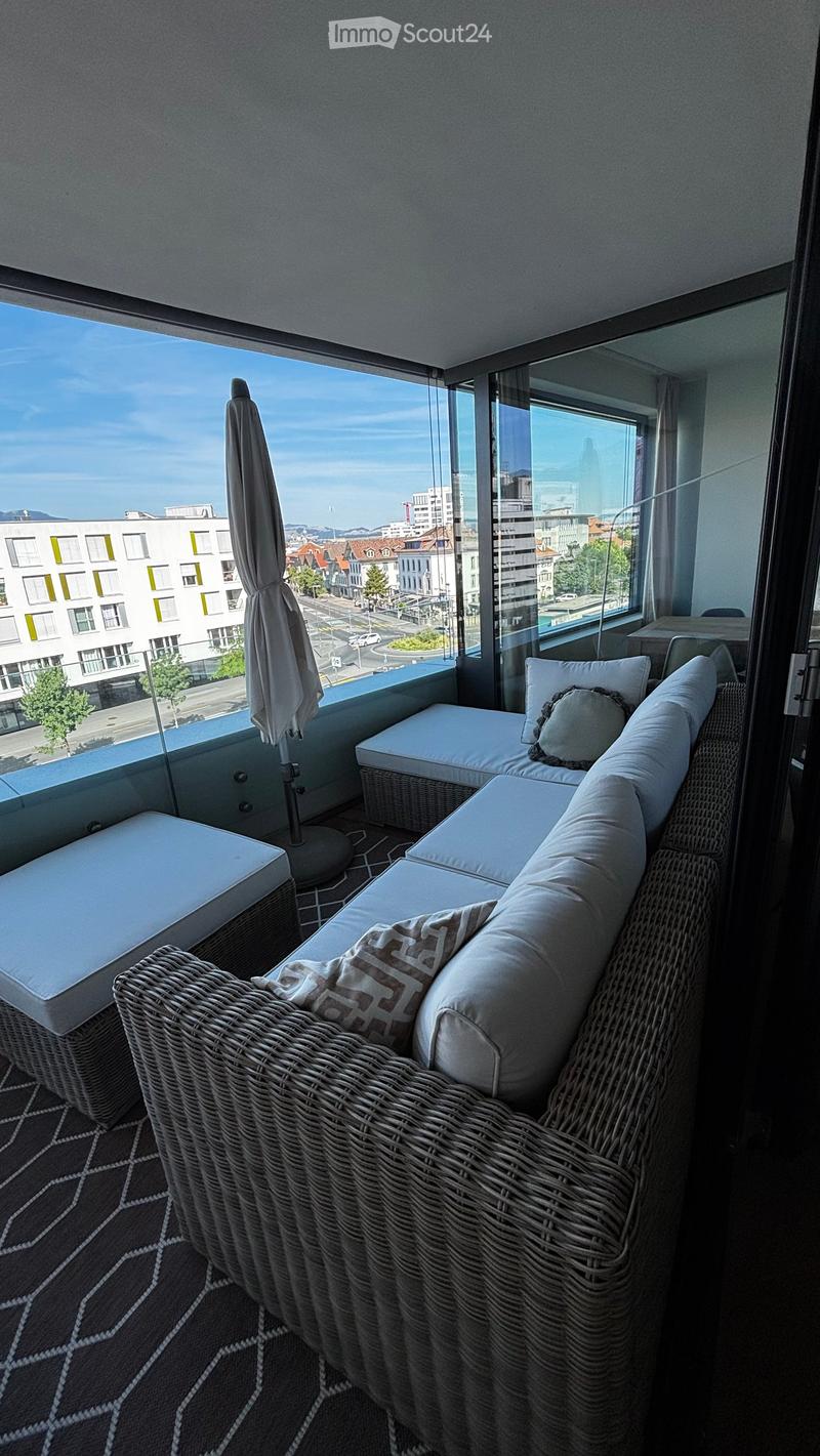 Balcony with wicker sofa, matching stools, and a folded umbrella, overlooking a cityscape.