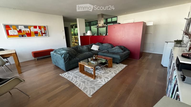 Modern living room with hardwood floors, grey sectional sofa, coffee table, wooden chair, red accent wall, and a colorful abstract painting.