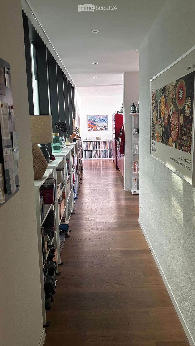 A long, narrow hallway with wooden flooring, white walls, and built-in shelving units on both sides. The left side features books, decorative items, and a lamp, while the right side has a large framed poster and a small shelf with items.