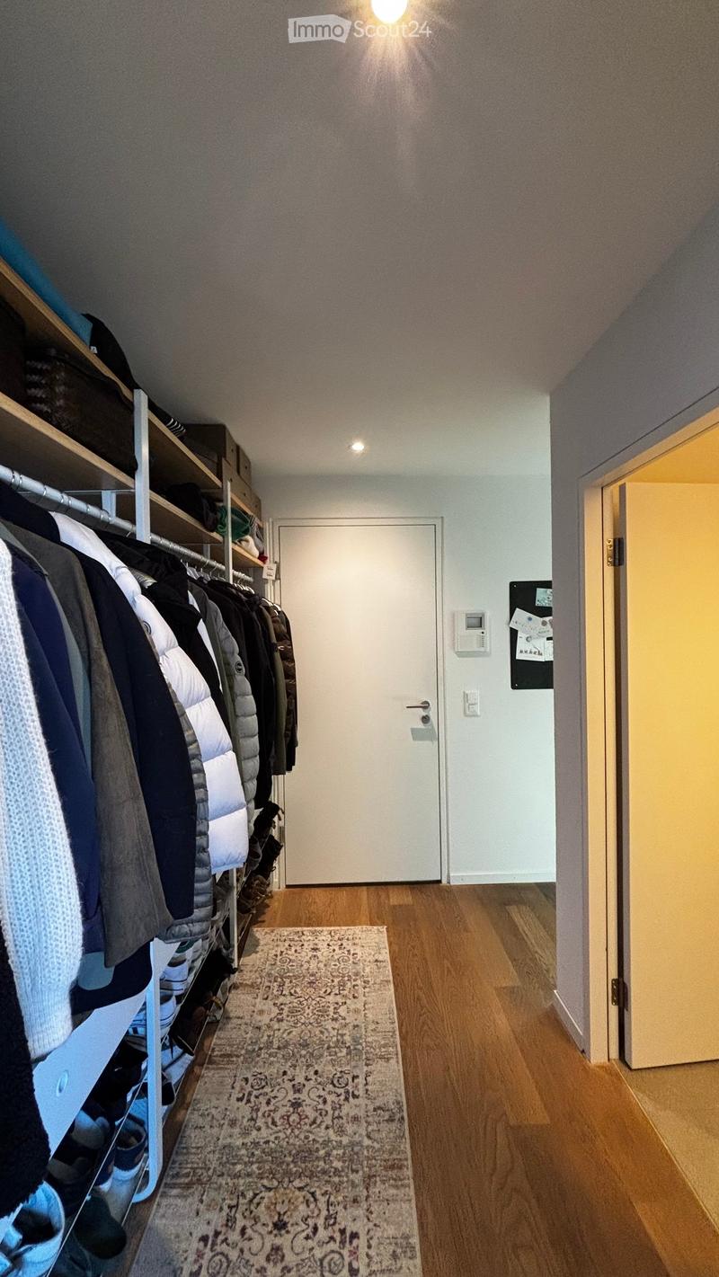 A well-lit hallway with wooden flooring, a patterned rug, and a closet with hanging clothes and shelves. A white door is visible at the end of the hallway.