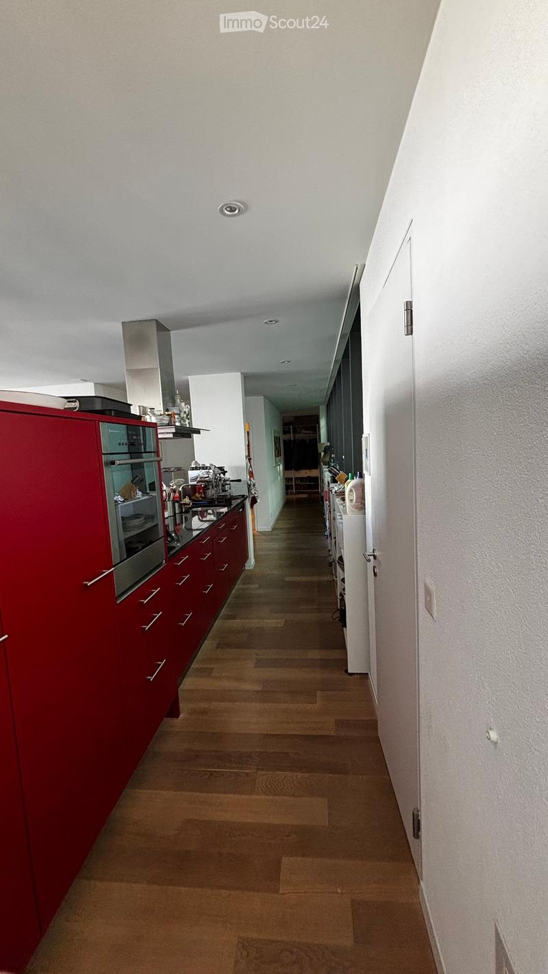 A narrow hallway with wooden flooring, white walls, and recessed ceiling lights. The hallway leads to a kitchen area with red cabinets and stainless steel appliances visible on the left side.