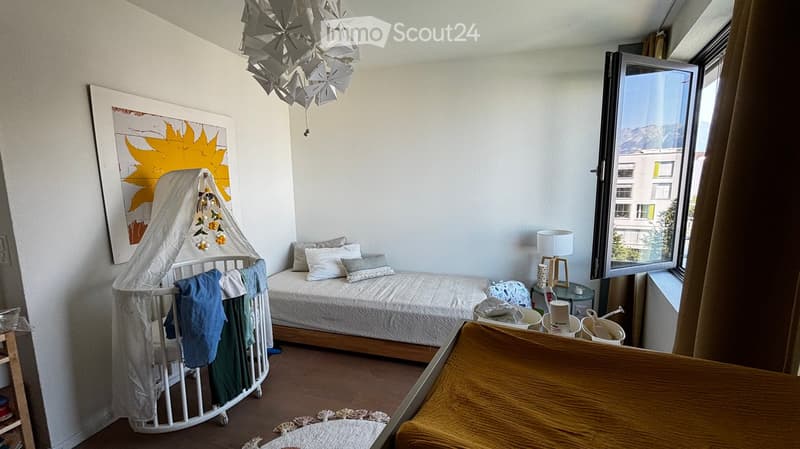 A cozy bedroom with a single bed, a crib, a small table with a lamp, and a large window with a view of mountains.