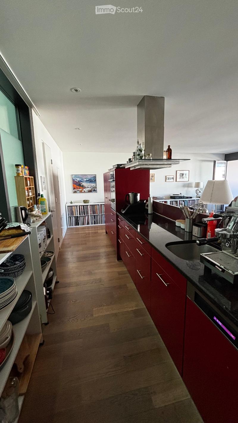 Modern kitchen with red cabinets, wooden flooring, and various appliances including a sink, oven, and coffee maker. Shelves hold dishes, bowls, and other kitchen items.