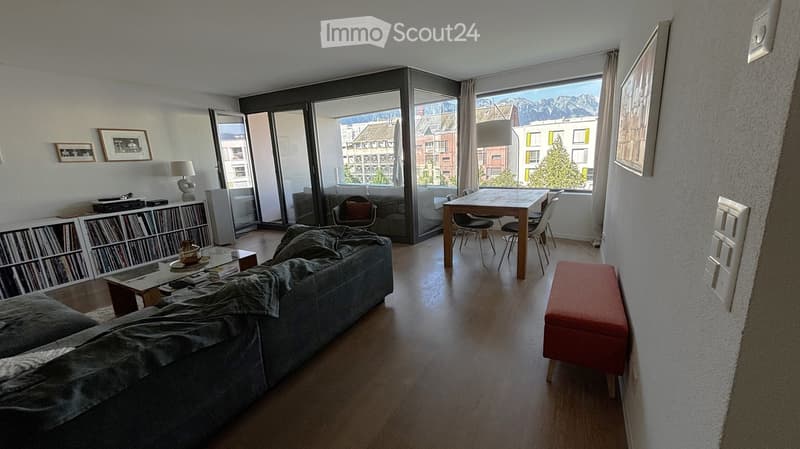 Modern living room with large windows offering a mountain view, featuring a grey sofa, coffee table, dining table with chairs, bookshelf, and a red ottoman.
