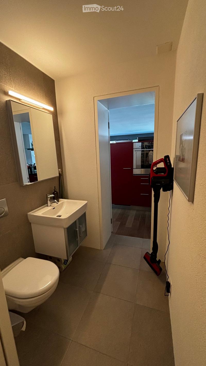 Modern bathroom with a toilet, sink, mirror, and a cabinet. A vacuum cleaner is plugged into the wall. The room has beige tiled flooring and light-colored walls.