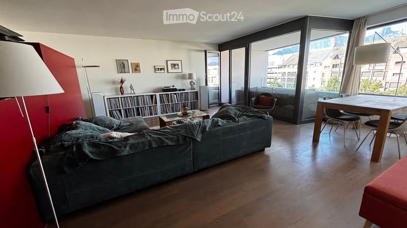 Modern living room with wooden flooring, large windows, a grey sofa, a coffee table, a dining table with chairs, and a bookshelf with books and decorative items.