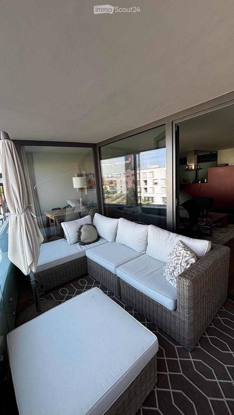 Balcony with white wicker sofa, matching chair, small table, and umbrella, overlooking cityscape.