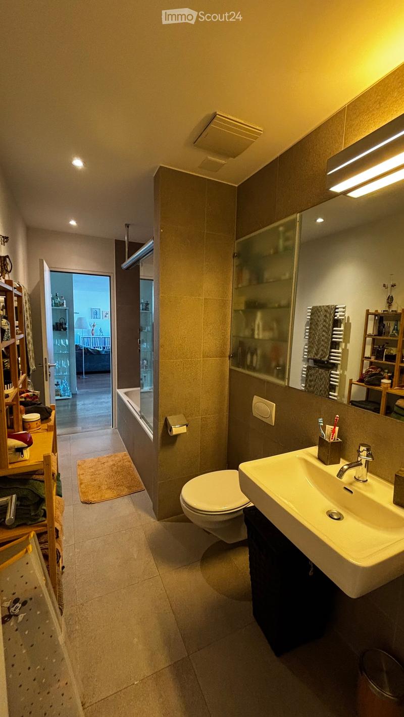 Modern bathroom with a white sink, toilet, and bathtub. Features a large mirror, towel rack, and shelves with various items. The room has beige tiles and warm lighting.