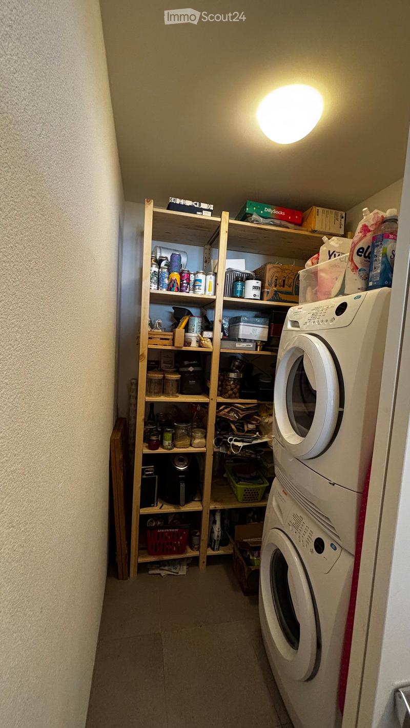 A small, well-lit laundry room with wooden shelves filled with various items, including jars, cans, and boxes. A white washing machine and dryer are positioned against the wall.