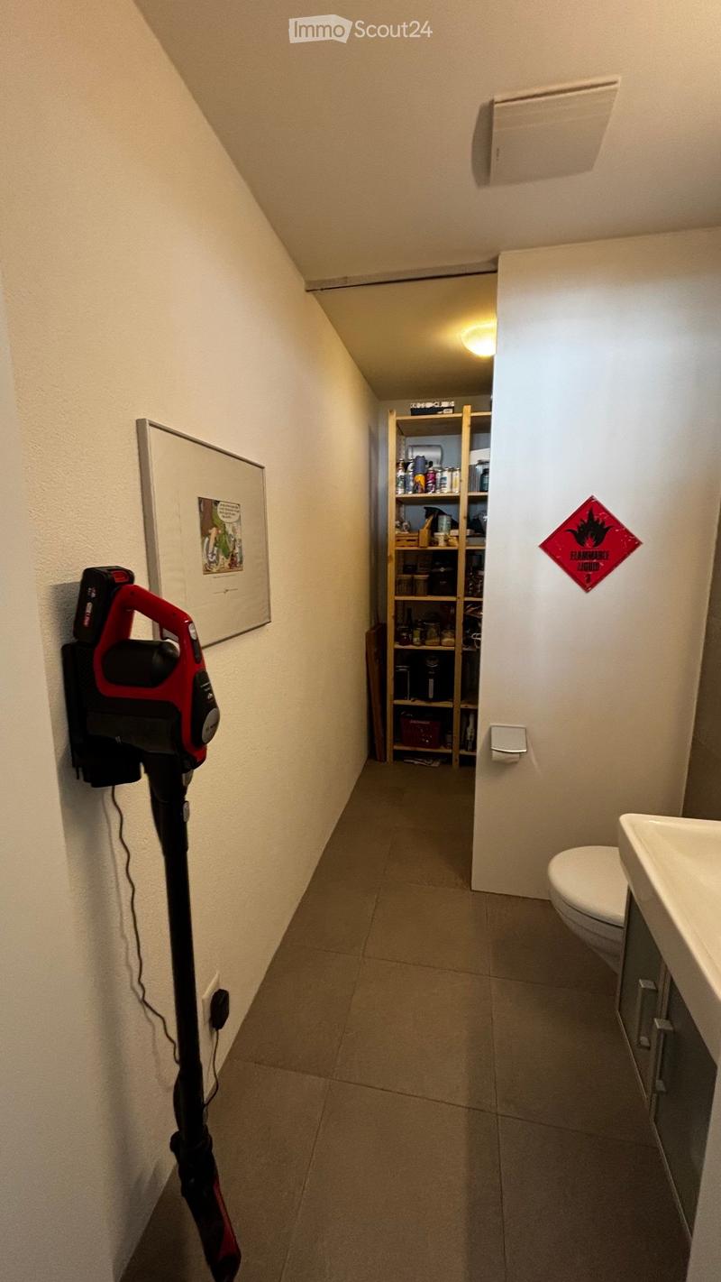 A narrow hallway with tiled flooring, white walls, and a small shelf unit at the end holding various items. A vacuum cleaner is mounted on the left wall, and a warning sign is visible on the right wall.