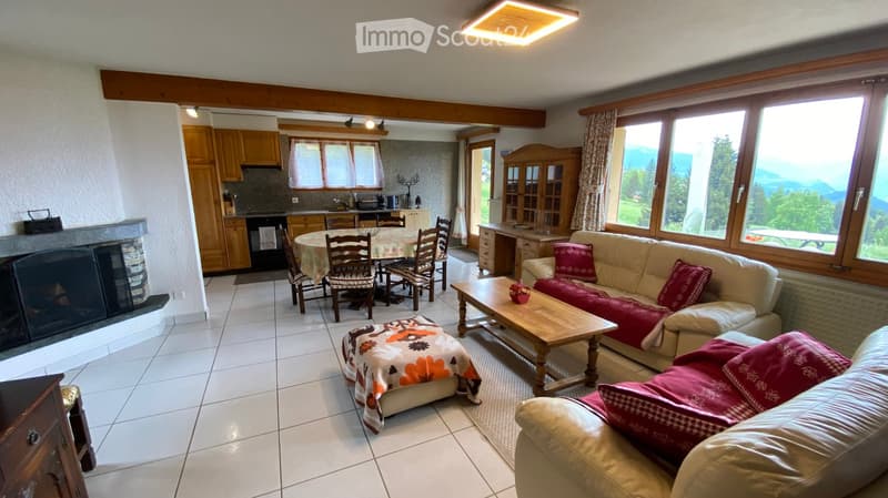 A spacious living room with a beige sofa, coffee table, ottoman, and fireplace. A dining area with a round table and chairs is visible, along with a kitchen area featuring cabinets, an oven, and a refrigerator. Large windows offer a scenic mountain view.