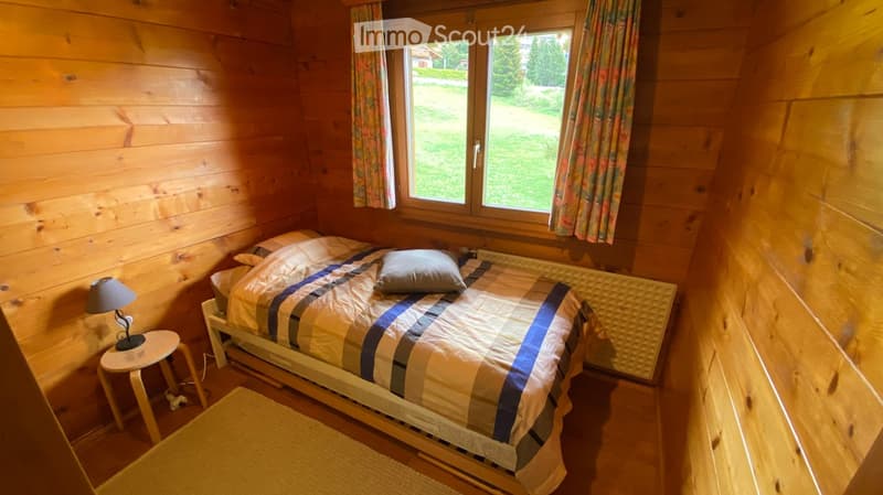 Cozy wooden bedroom with a single bed, nightstand, lamp, and rug. Large window with floral curtains offers a view of the garden.