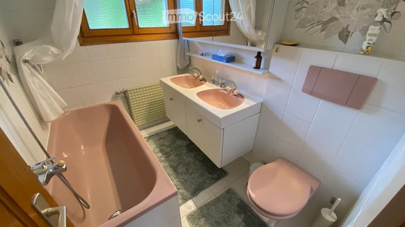 Bathroom with pink bathtub, double sink, toilet, mirror, and white tiled walls.