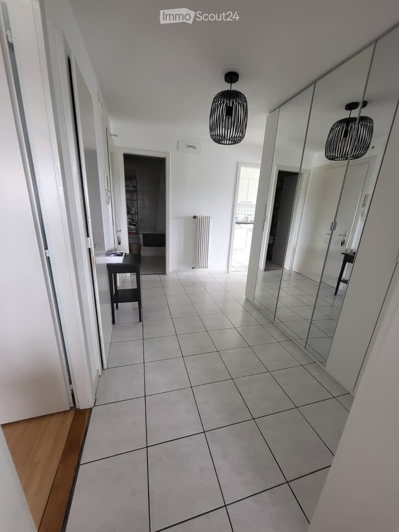 Clean, modern hallway with white tiled flooring, white walls, and a large mirror on one side. Features include a small black table, a hanging woven light fixture, and visible doorways leading to other rooms.