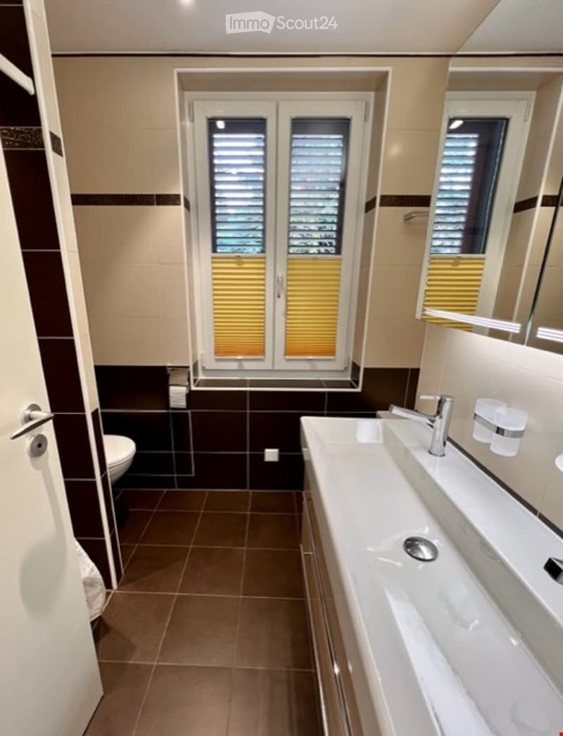 Modern design bathroom with brown tiles, white sink, silver faucet, toilet, and window with yellow blinds.