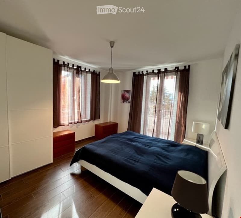 wooden floor, large bed, brown curtains, lamp on nightstand, white cabinet
