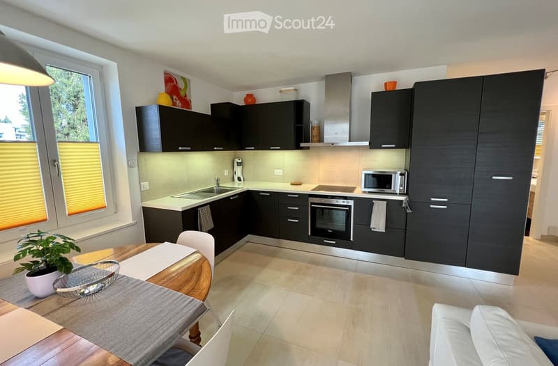 modern kitchen with wooden table, black cabinets, built-in oven, microwave, dishwasher, washing machine, window with yellow blinds, potted plant
