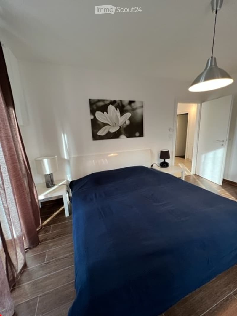 The bedroom is furnished with a bed and a night table with a lamp on both sides. The wall is white with a black and white framed picture. The bed has a dark blue duvet. There is a white lamp over the bed.