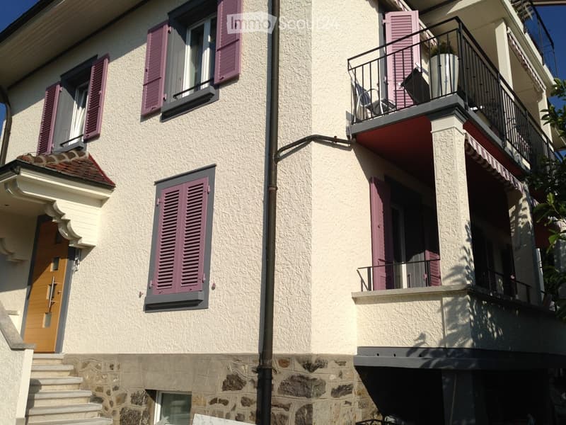 Building exterior with three floors. Red roof, white walls, red shutters on the windows. Two balconies, one with an awning. A chair and potted plant on the balcony.