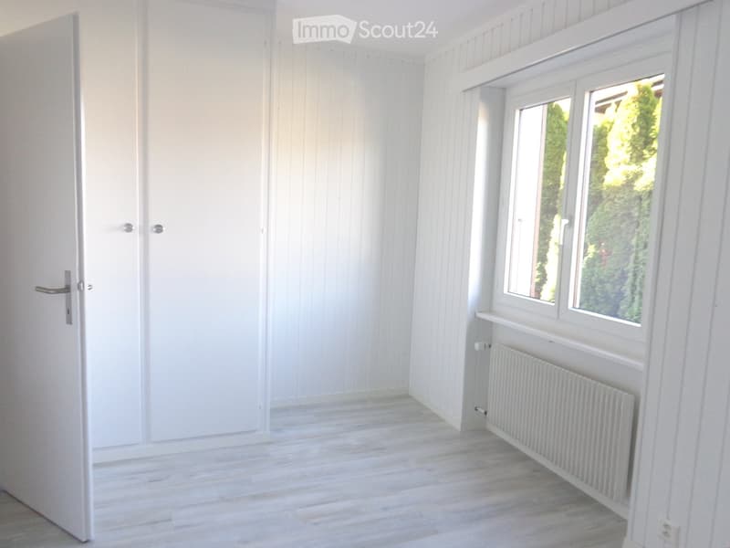 Empty room with white paneled walls, light wooden flooring, a white door, and a window with a radiator below it.