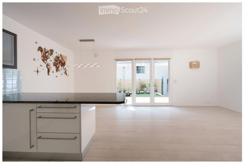 Open-plan space with white walls, light wooden flooring, and large sliding glass doors leading to an outdoor area. Features a modern kitchen island with white cabinets and a black countertop, a world map wall decoration, and a wall-mounted light fixture.