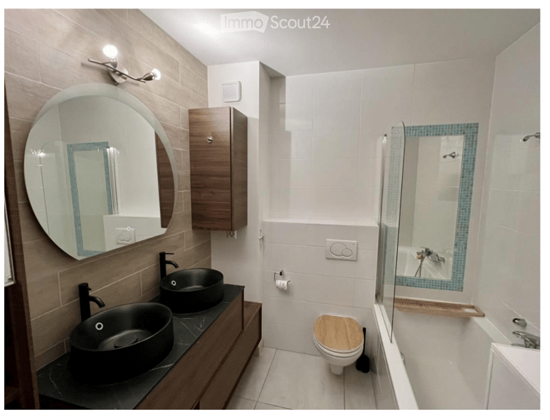Modern bathroom with two black sinks, a round mirror, a wooden toilet seat, and a glass-enclosed shower with blue tile accents.