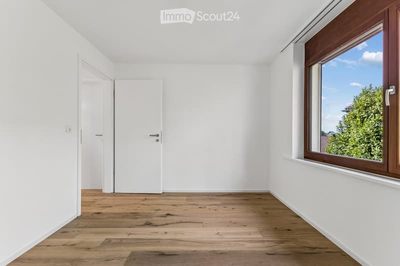 Empty room with white walls, wooden flooring, and a large window with wooden frame showing a view of greenery outside. There is a white door on the left side.