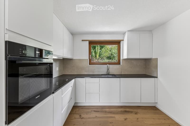 Modern kitchen with white cabinets, black countertops, a built-in oven, and a window overlooking greenery.