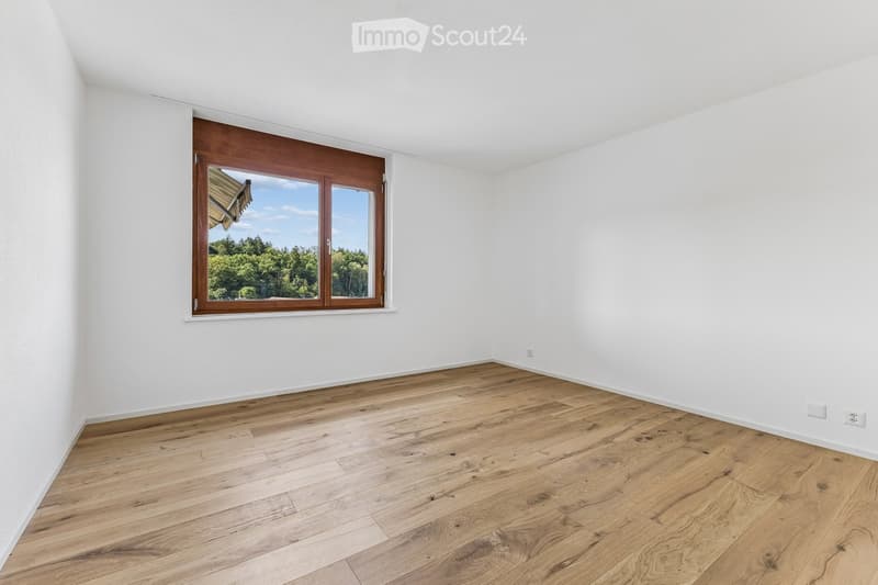 Empty room with light wooden flooring and white walls, featuring a large window with wooden frame and glass panes, offering a view of greenery outside.