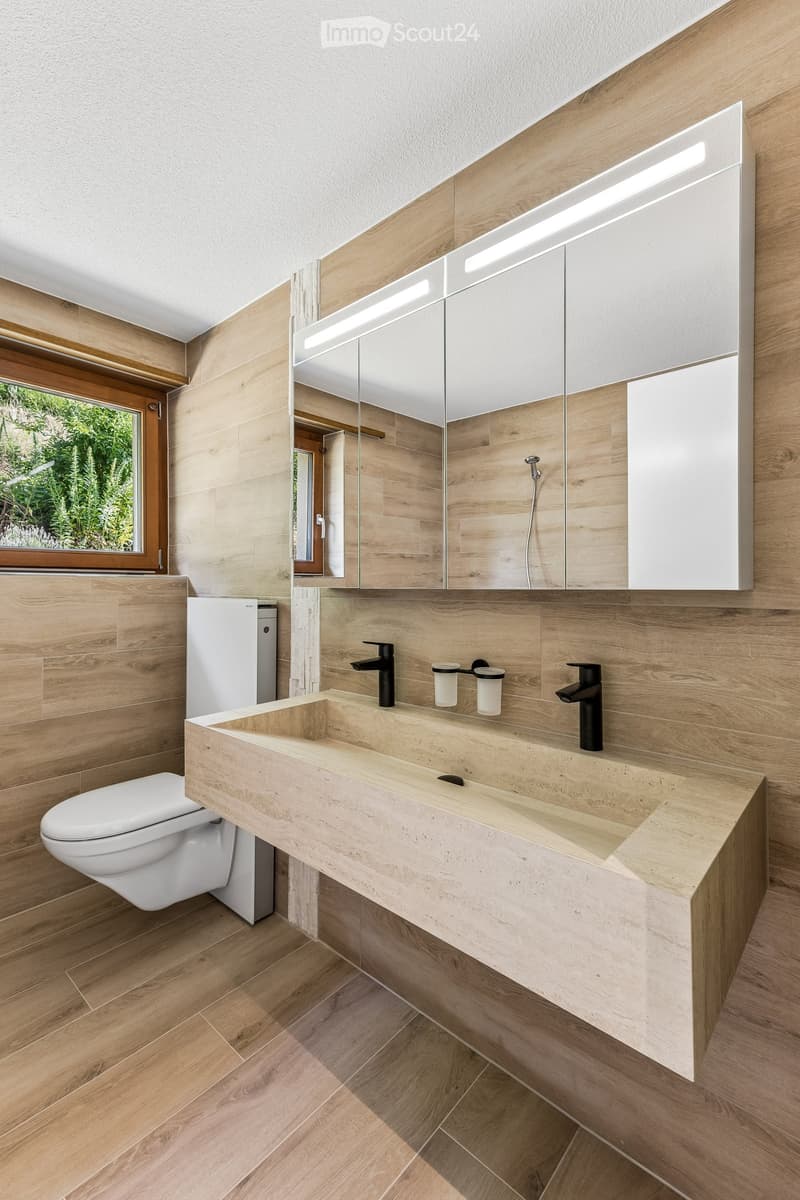 Modern bathroom with wooden walls, a large sink with two faucets, a mirror cabinet, and a toilet. The space features wooden flooring and a window with a view of greenery.