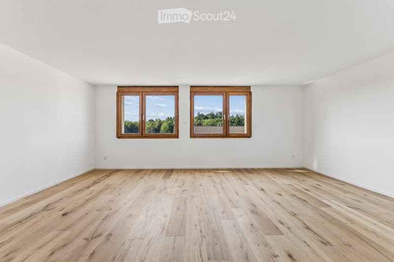 Empty room with light wooden flooring and white walls, featuring two large windows with wooden frames offering a view of greenery outside.