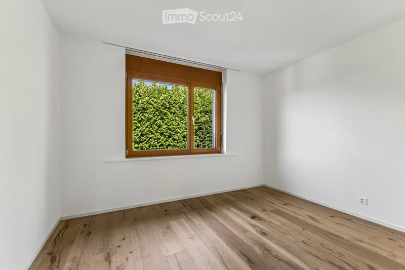 Empty room with white walls, wooden flooring, and a large window with wooden frame showing greenery outside.