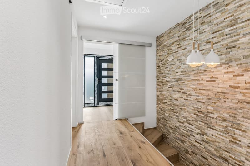 Modern hallway with wooden flooring, white walls, and a textured stone accent wall. Features pendant lights and a glass sliding door leading to another room.