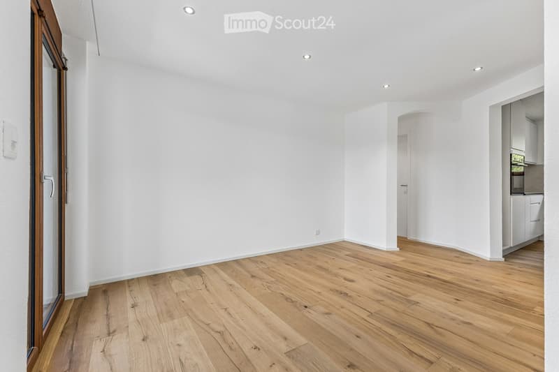 Empty room with white walls, wooden flooring, and recessed ceiling lights. Features a large window with a wooden frame and a doorway leading to another room.