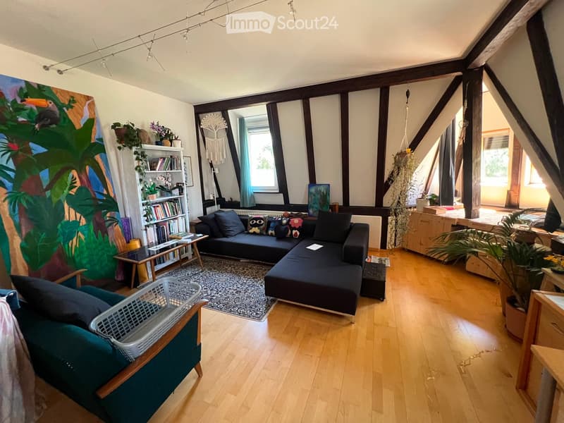 A cozy living room with wooden flooring, a black sectional sofa, a green armchair, a coffee table, a bookshelf, and various decorative items including plants and artwork.