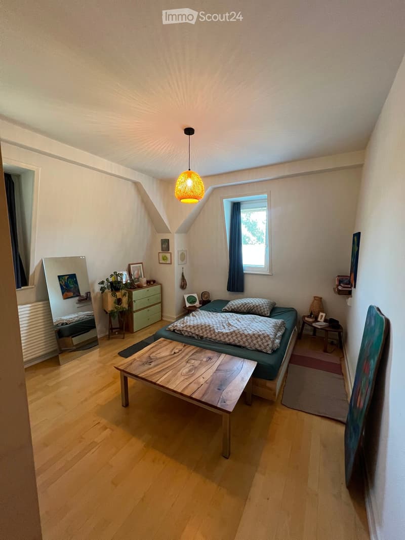 Cozy bedroom with wooden flooring, a bed with patterned bedding, a wooden table, a mirror, a small cabinet, and a hanging lamp.