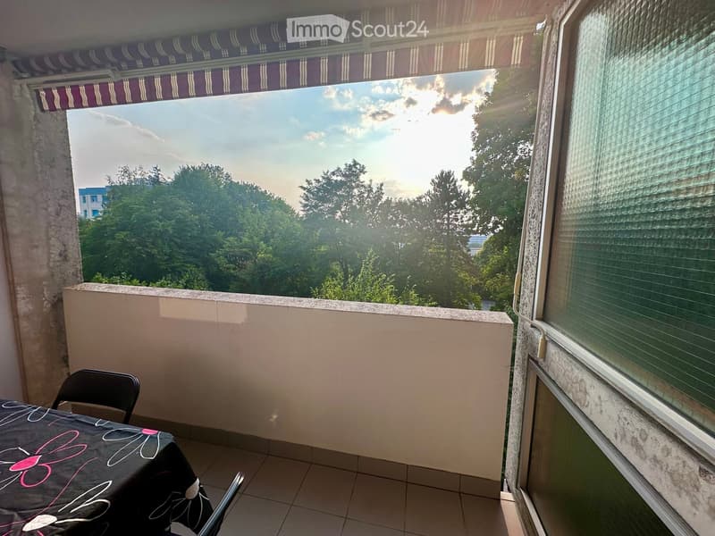 Balcony with tiled floor, table, and chair, overlooking a tree-filled area with a building in the distance.