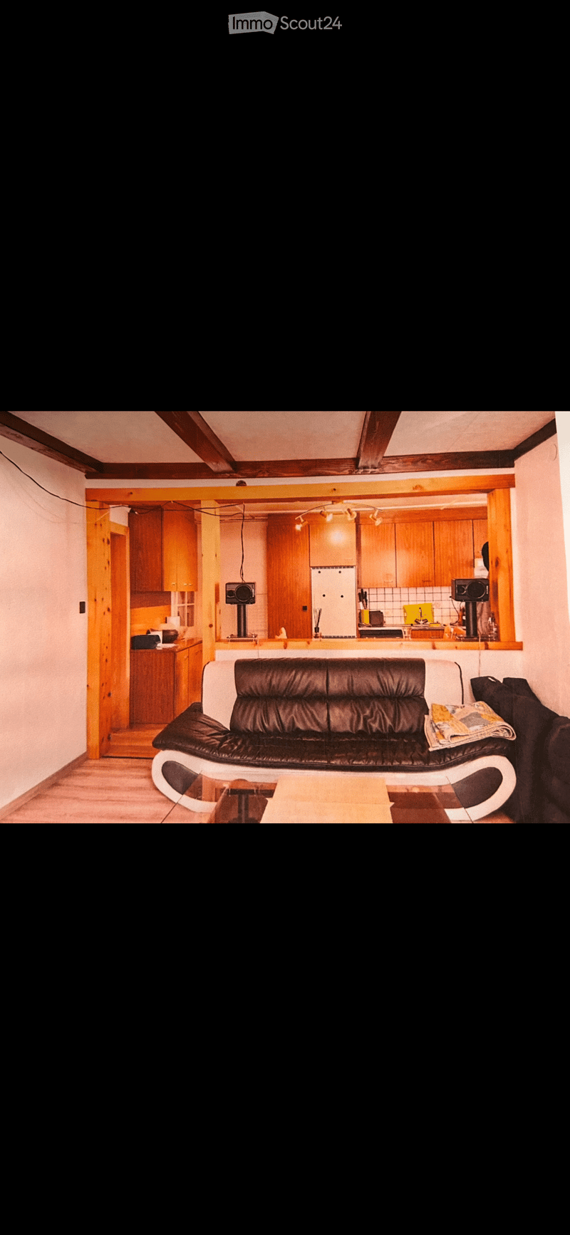 Open plan living room and kitchen, wooden floor, wooden beams, black leather sofa, wooden coffee table, wooden cabinets, white refrigerator, kitchen appliances