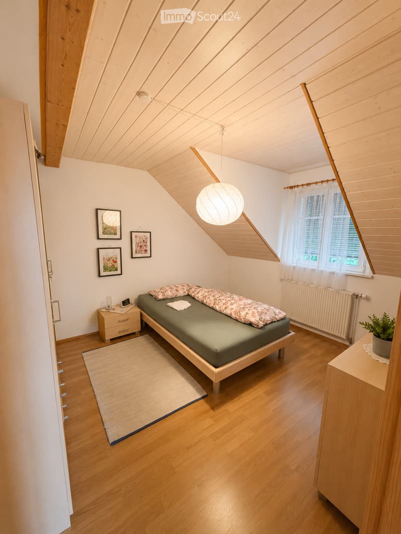 Cozy attic bedroom with wooden floors, slanted wooden ceiling, bed with green mattress and patterned pillows, nightstand with lamp, framed pictures on wall, potted plant on dresser, and large window with white curtains.