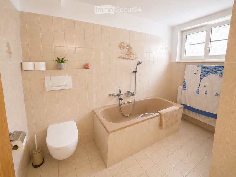 Bathroom with beige tiles, a bathtub, toilet, towel, and a small plant on the shelf.