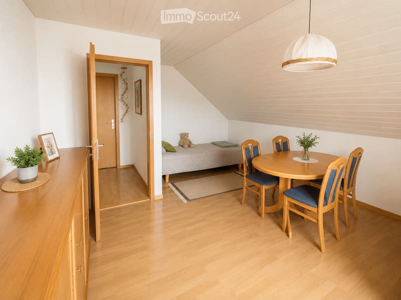 Bright, wooden-floored room with a dining area featuring a round table and four chairs, a small sofa with a teddy bear and pillow, a cabinet with a plant, and a picture frame on the wall.