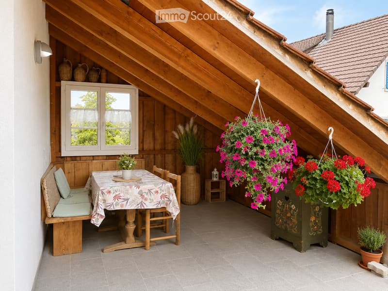 Cozy wooden balcony with slanted wooden ceiling, tiled floor, and large window overlooking greenery. Features a small dining table with chairs, a bench, hanging flower baskets, and decorative vases.