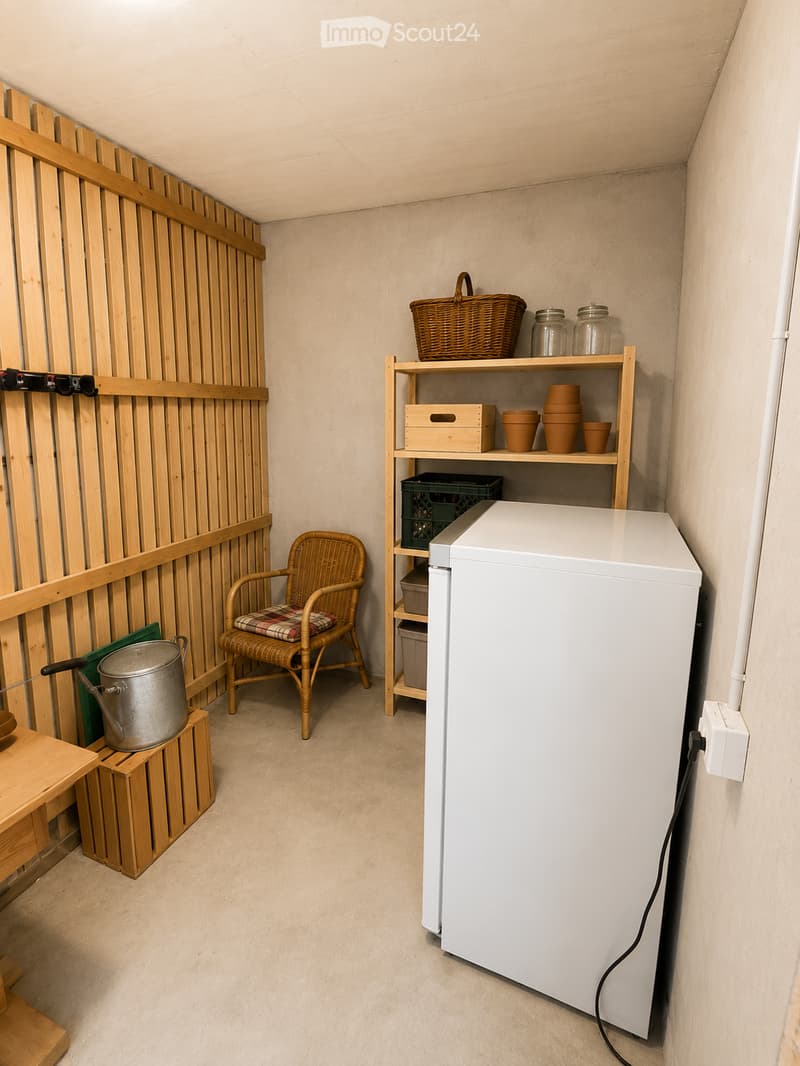 A small, furnished room with a refrigerator, a wicker chair with a cushion, a wooden shelf holding jars and cups, a metal pot on a wooden crate, and a woven basket on top of the shelf.