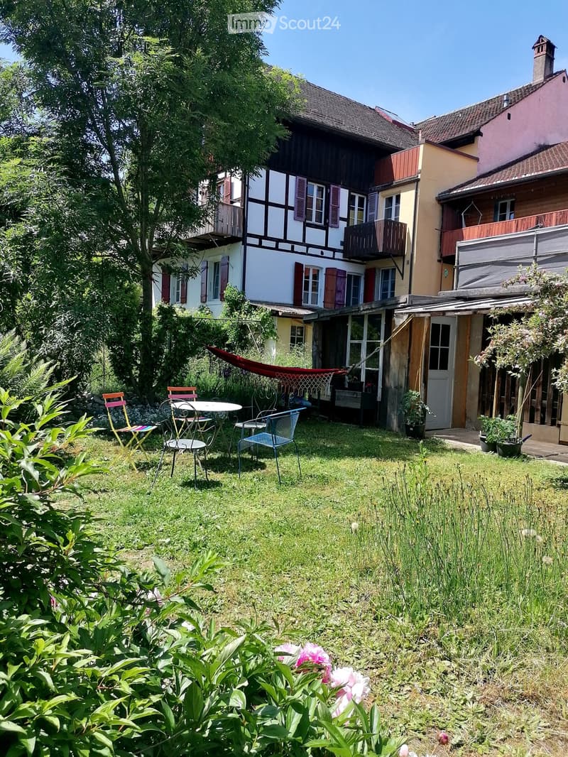 Garden area with a table, chairs, hammock, and surrounding greenery near a multi-story house.